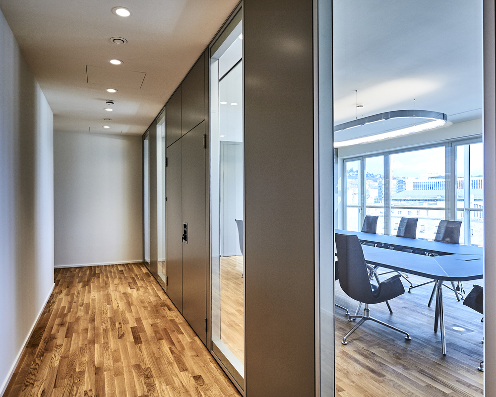 Corridor in a modern office with strip parquet flooring, illuminated by small round recessed spotlights, BE2 luminaires. Through a glass front, you can see into an adjacent meeting room with a table, chairs and rectangular SOFT SQUARE ring luminaire.