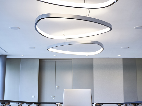 Three polished aluminium ring luminaires in the shape of a rounded triangle (SOFT DELTA) are suspended from the ceiling in a row with different orientations, underneath which stands a light-coloured chair by WALTER KNOLL on brown parquet flooring. | © Fotografie SUL – Sven Lorenz