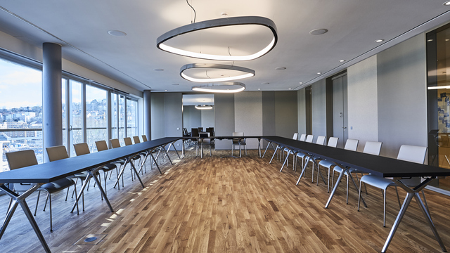 Konferenzraum mit U-Bestuhlung in Brauntönen, Möbel von WALTER KNOLL, in der Mitte des Raumes sind drei große Ringleuchten in Wankelform, SOFT DELTA, aus poliertem Aluminium abgependelt,  graue Wände an den Seiten, durch eine Tür im Hintergrund kann man in einen weiteren Besprechungsraum blicken. | © Fotografie SUL – Sven Lorenz