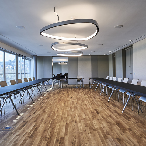 Konferenzraum mit U-Bestuhlung in Brauntönen, Möbel von WALTER KNOLL, in der Mitte des Raumes sind drei große Ringleuchten in Wankelform, SOFT DELTA, aus poliertem Aluminium abgependelt,  graue Wände an den Seiten, durch eine Tür im Hintergrund kann man in einen weiteren Besprechungsraum blicken.