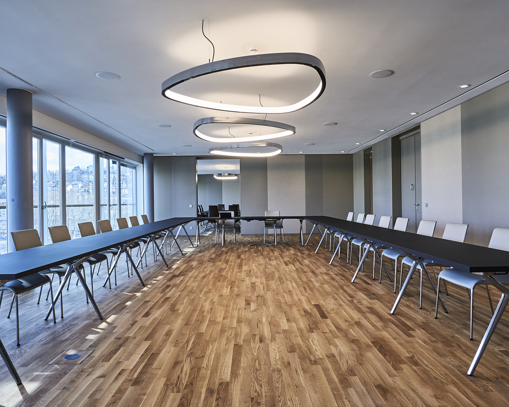 Konferenzraum Beleuchtung Konferenzraum mit U-Bestuhlung in Brauntönen, Möbel von WALTER KNOLL, in der Mitte des Raumes sind drei große Ringleuchten in Wankelform, SOFT DELTA, aus poliertem Aluminium abgependelt, graue Wände an den Seiten, durch eine Tür im Hintergrund kann man in einen weiteren Besprechungsraum blicken.