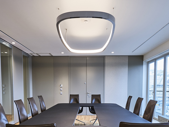 Blick von der kurzen Seite eines rechteckigen Besprechungstisches schräg nach oben an die Decke, von der eine Ringleuchte in Form eines abgerundeten Rechtecks abgependelt ist. Außen in poliertem Aluminium innen mit einem Diffusor, leuchtet sie nach innen und dabei erhellt sie sowohl die Decke, als auch den Tisch. | © Fotografie SUL – Sven Lorenz