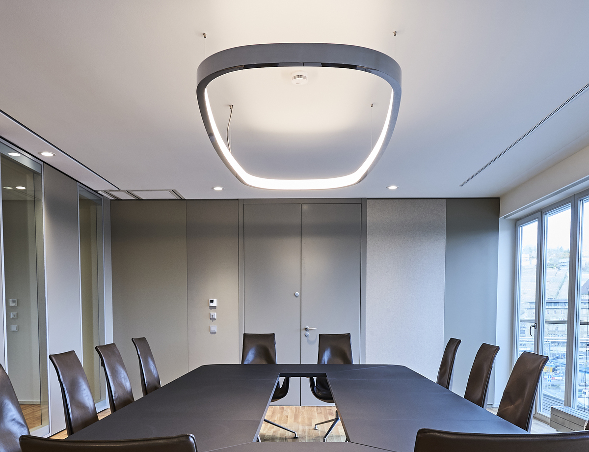 Blick von der kurzen Seite eines rechteckigen Besprechungstisches schräg nach oben an die Decke, von der eine Ringleuchte in Form eines abgerundeten Rechtecks abgependelt ist. Außen in poliertem Aluminium innen mit einem Diffusor, leuchtet sie nach innen und dabei erhellt sie sowohl die Decke, als auch den Tisch.