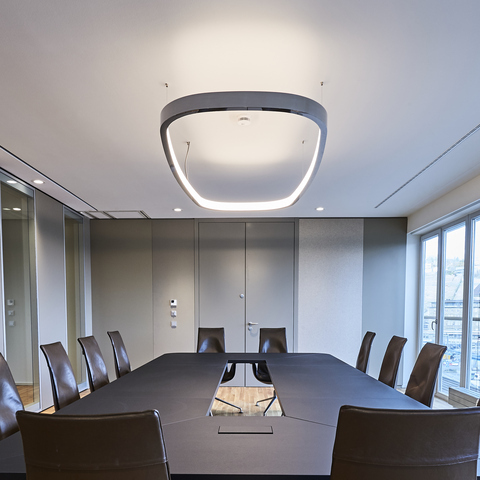 Blick von der kurzen Seite eines rechteckigen Besprechungstisches schräg nach oben an die Decke, von der eine Ringleuchte in Form eines abgerundeten Rechtecks abgependelt ist. Außen in poliertem Aluminium innen mit einem Diffusor, leuchtet sie nach innen und dabei erhellt sie sowohl die Decke, als auch den Tisch.
