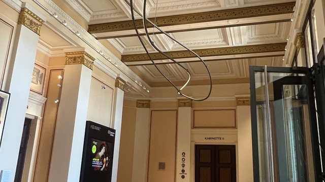 Saal mit Sitzgruppe und geschwungener Lichtskulptur an der Decke zwischen Säulen | © Ulrich Sattler