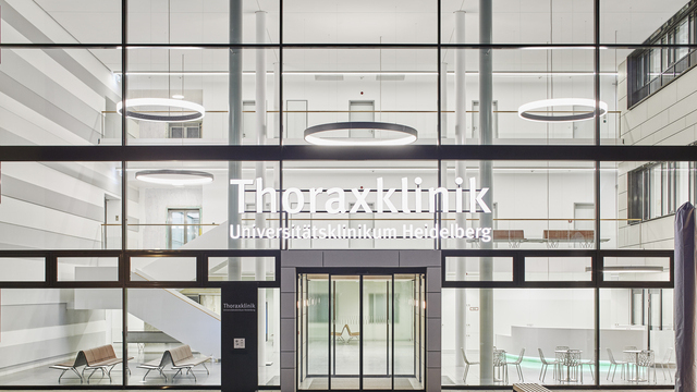 Landscape: Exterior view through glass front onto the entrance hall of the thorax clinic with columns and large ring lights suspended on different levels | © Dietmar Strauß / Besigheim