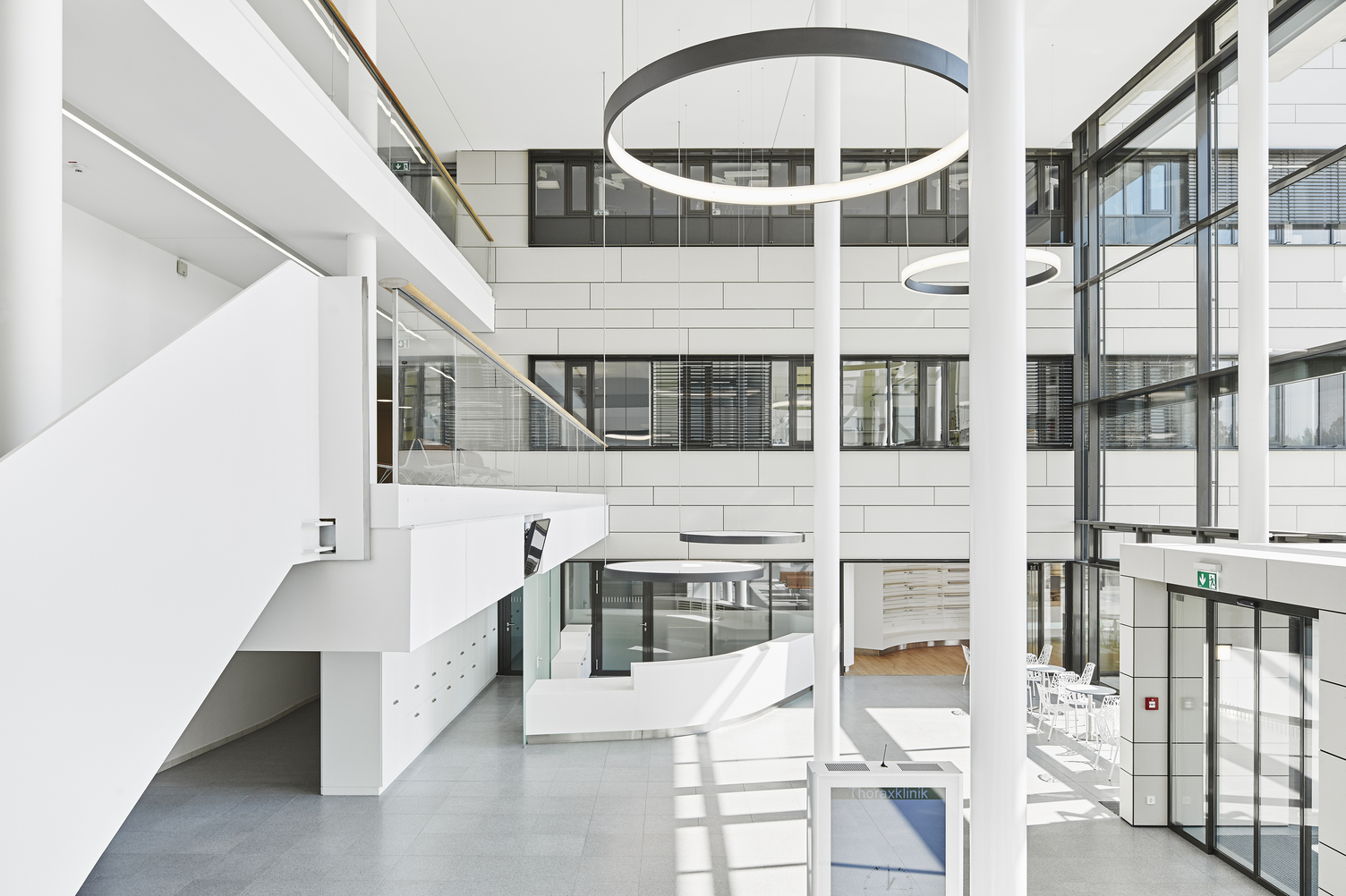 Landscape: High white entrance hall of the Thorax Clinic with a view of the reception desk, above which two round luminaires with acoustic foil are suspended to provide privacy, in the room columns and 2 large black ring luminaires radiating inwards (CIRCOLO I) and 2 ring luminaires (CIRCOLO INVERSE) suspended in the room radiating outwards.