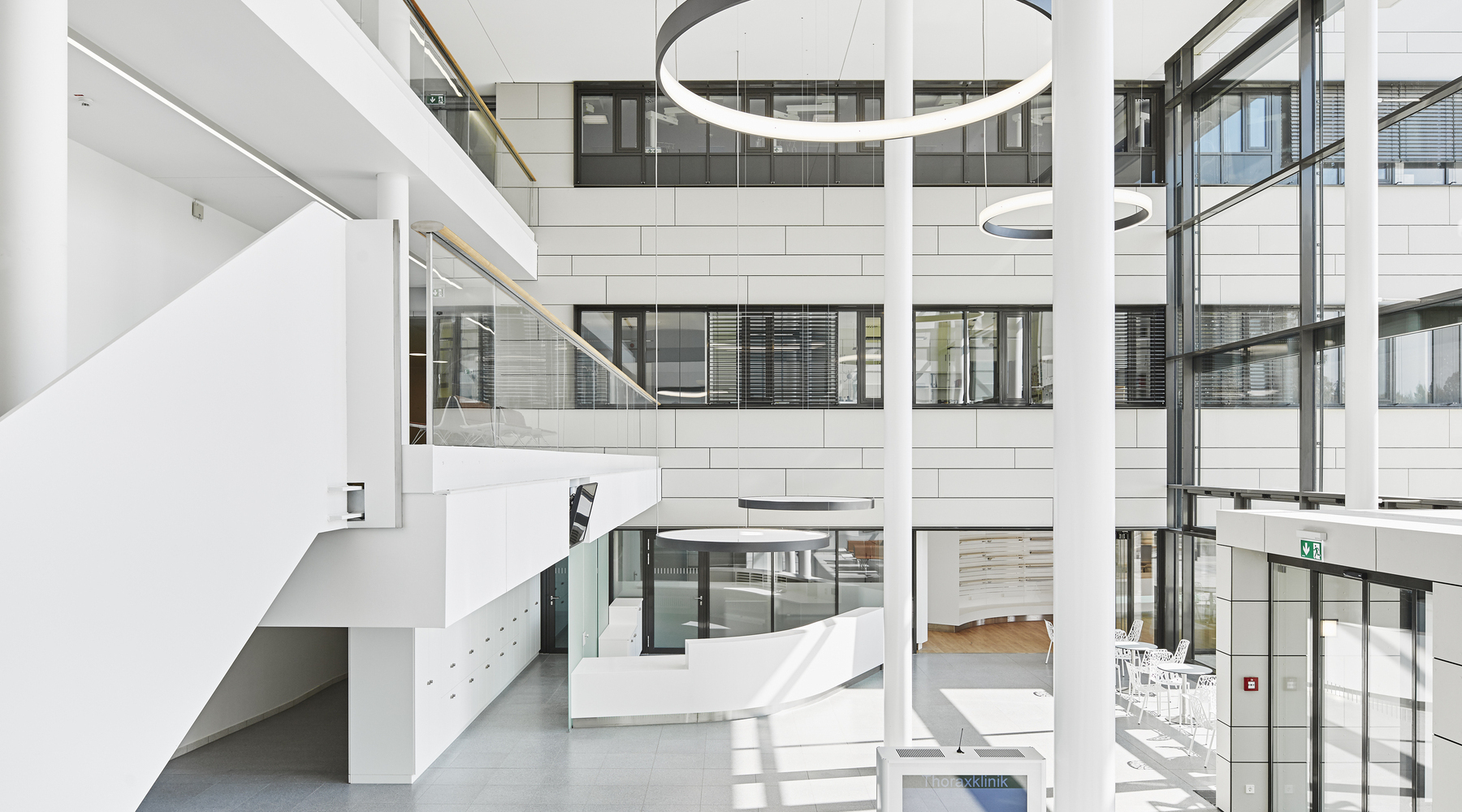 Empfangstheke mit Akustikleuchte CIRCOLO L+A - Eingangshalle & Treppenhaus Thoraxklinik mit CIRCOLO Ringleuchten | © Dietmar Strauß / Besigheim Querbild: Hohe weiße Eingangshalle der Thoraxklinik mit Blick auf die Empfangstheke, über der zwei runde Leuchten mit Akustikfolie zum Sichtschutz hängen, im Raum Säulen und 2 große schwarze Ringleuchten nach innenstrahlend (CIRCOLO I) und 2 Ringleuchten (CIRCOLO INVERSE) nach außenstrahlend im Raum abgehangen. | © Dietmar Strauß / Besigheim