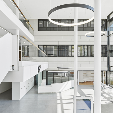 Landscape: High white entrance hall of the Thorax Clinic with a view of the reception desk, above which two round luminaires with acoustic foil are suspended to provide privacy, in the room columns and 2 large black ring luminaires radiating inwards (CIRCOLO I) and 2 ring luminaires (CIRCOLO INVERSE) suspended in the room radiating outwards.
