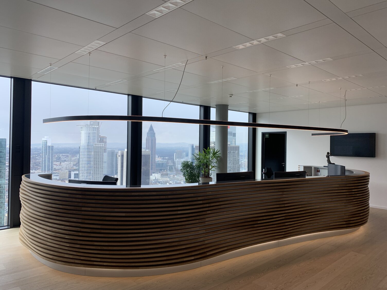 Maßgeschneiderte Pendelleuchte DUETTO über Empfangstresen | © TRITON Großer Empfangstresen vor einer großen Fensterfront im Bürogebäude von TRITON im Omni Tower Frankfurt. In der Form ist die lange Empfangstheke geschwungen und scheint optisch von außen mehrrippig aus dunklen Holz aufgebaut, darüber ist eine lineare schwarze Pendelleuchte DUETTO in gleicher geschwungener Form abgehangen. Maßgeschneiderte Sonderleuchte für die Kundentheke. | © TRITON