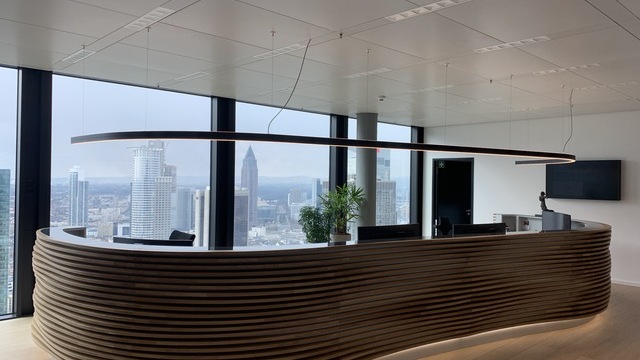 Large reception counter in front of a large window front in the TRITON office building in the Omni Tower Frankfurt. The long reception counter is curved in shape and appears to have a multi-ribbed structure made of dark wood from the outside, with a linear black DUETTO pendant luminaire suspended above it in the same curved shape. Customised special luminaire for the customer counter. | © TRITON