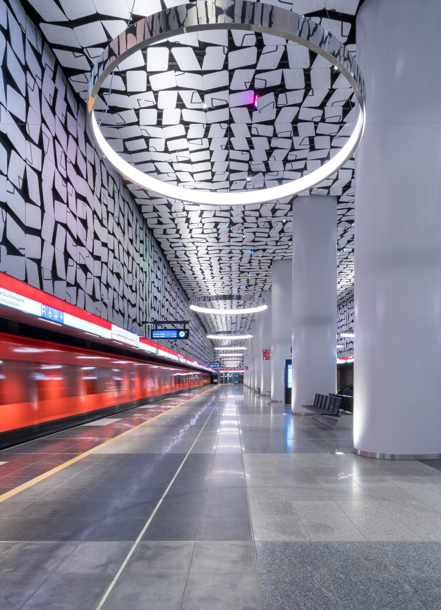 Durch das Bild fahrende orangene U-Bahn, die U-Bahnstation besteht an den Wänden und Decken aus grafischen Mustern, die sich in den von der Decke abgependelten runden Ringleuchten (CIRCOLO) spiegeln.