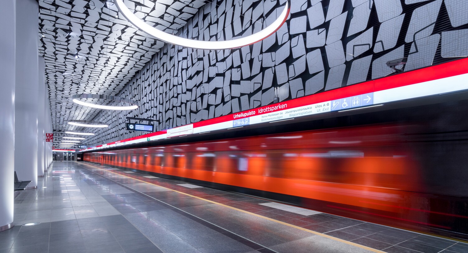 Durch das Bild fahrende orangene U-Bahn, die U-Bahnstation besteht an den Wänden und Decken aus grafischen Mustern, die sich in den von der Decke abgependelten runden Ringleuchten (CIRCOLO) spiegeln.