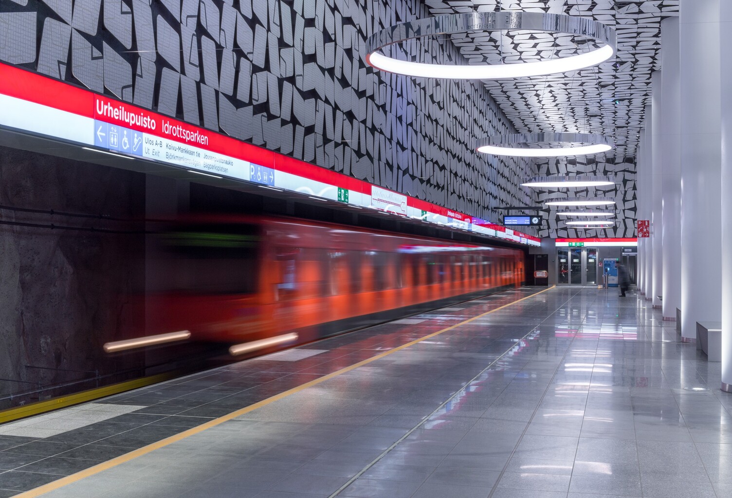 Orange underground train passing through the image, the underground station consists of graphic patterns on the walls and ceilings, which are reflected in the round CIRCOLO ring lights suspended from the ceiling.