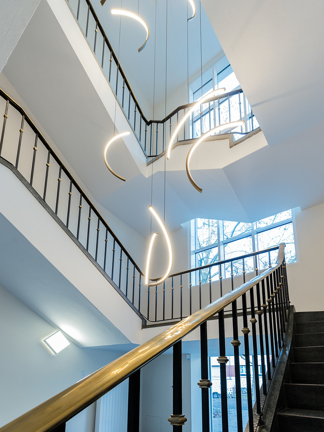 Kronleuchter/ Lichtobjekt im Treppenauge eines Firmengebäudes der VCC Group, bestehend aus Kreis-Segmenten der SLIM Ringleuchten nach innen leuchtend und dem Leuchtenprofil aus Aluminium in gold. | © eydos GmbH