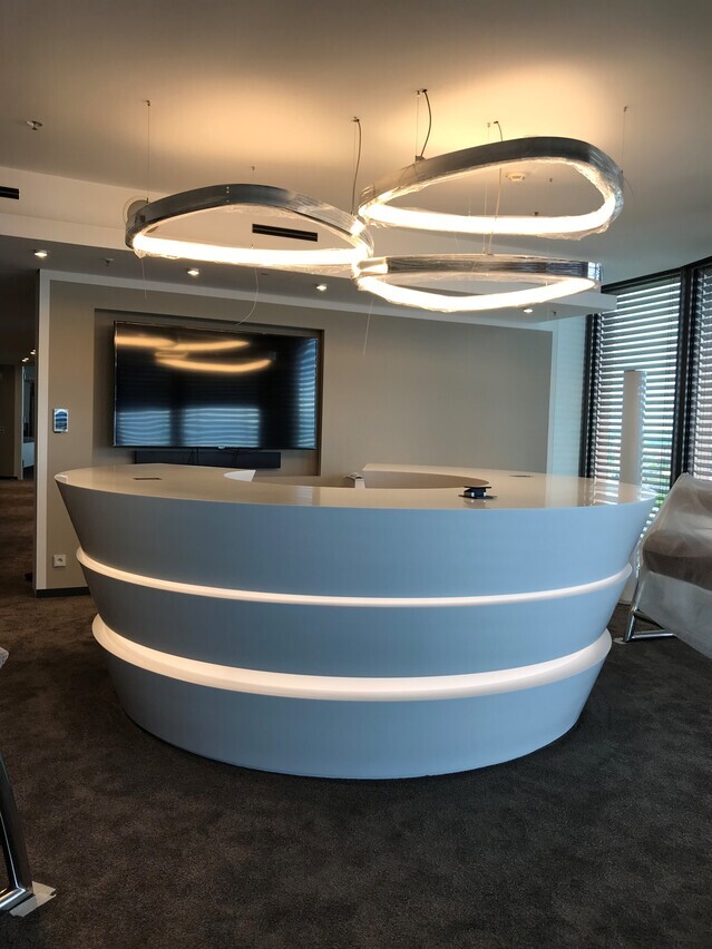 Three large-format aluminium ring lights in the shape of a rounded triangle, SOFT DELTA, suspended above a round white reception desk in an office building.
