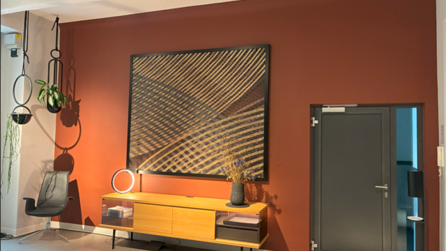 On the right of the picture is a door, on the left a red wall in front of which stands a wooden sideboard with a small black vertical light ring, SLIM TABLE, on top of it. Above it hangs a graphic picture. In the left-hand corner is a leather armchair by Walter Knoll.