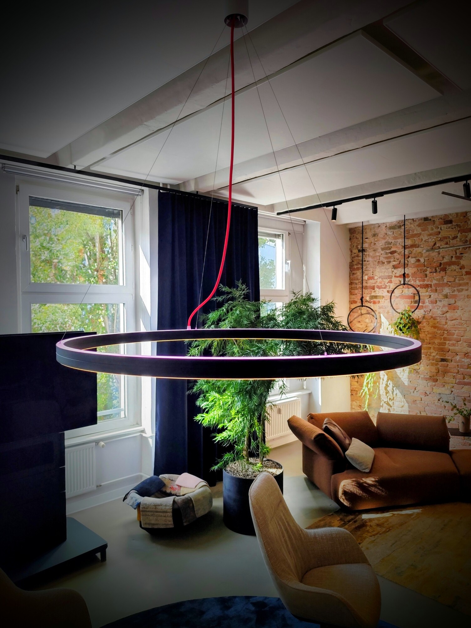 RGB Ringleuchte Doppio - direkt und indirekt leuchtend Runde Pendelleuchte Doppio, die nach unten weiß und nach oben rosa strahlt, abgependelt an mehreren dünnen Stahlseilen und Stromzuführung über ein rotes Textilstromkabel. Im Hintergrund ist ein Fernsehbildschirm und ein Sessel zu sehen.