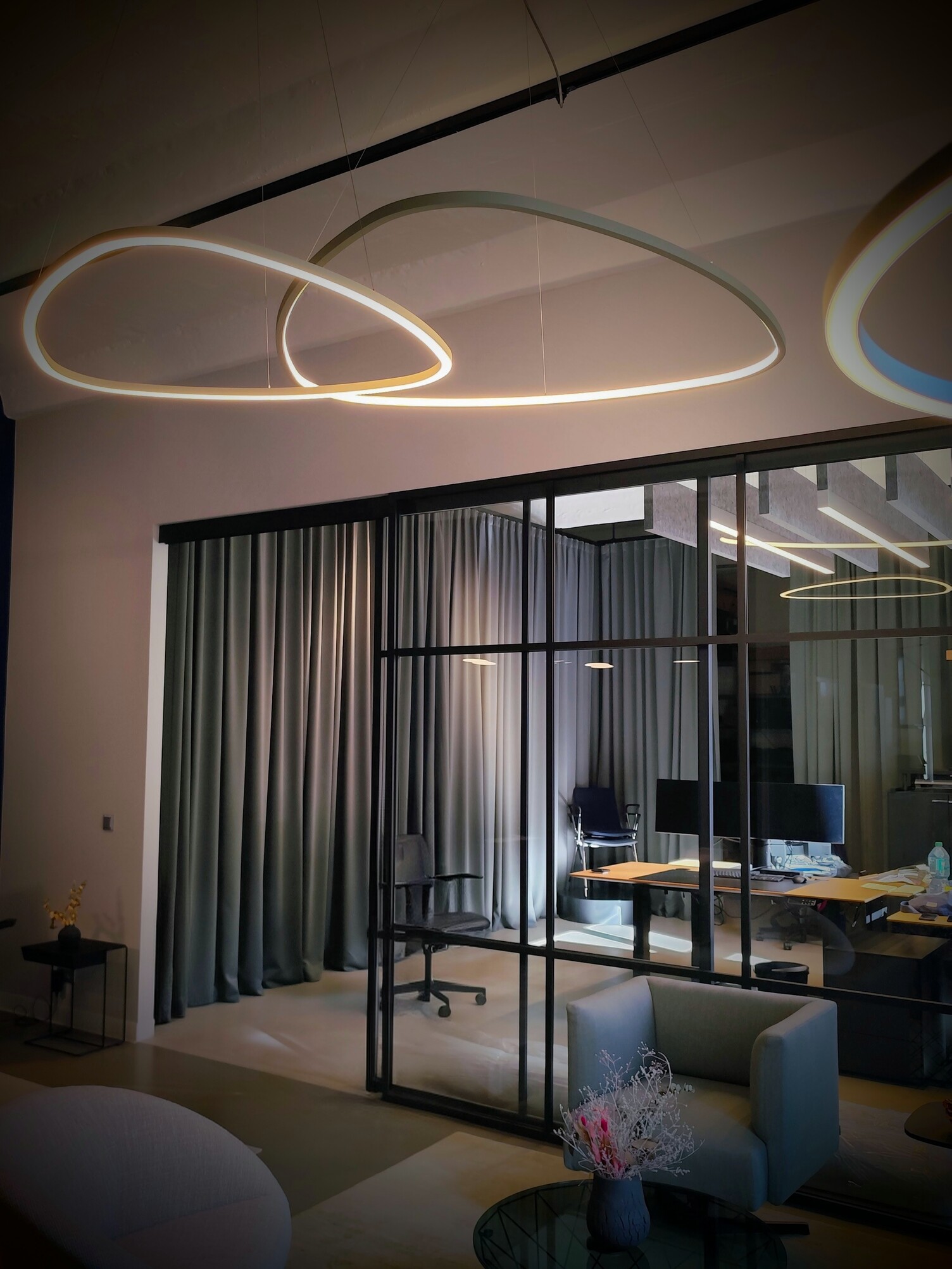 Dreiecksleuchten über Loungebereich Loungebereich mit dünnen dreieckigen Ringleuchten, SLIM DELTA, die darüber abgependelt sind. Im Hintergrund eine Glaswand und offene Durchgangstür, die in einen Bürobereich führt. In dem hinteren Raum sind eine Reihe Baffelleuchten und reine Akustikbaffeln über den Tischen aufgereiht abgehangen.