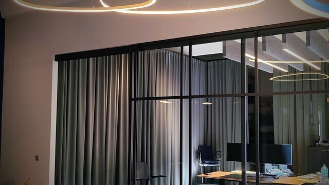 Lounge area with thin triangular ring lights, SLIM DELTA, suspended above. In the background, a glass wall and open doorway leading to an office area. In the rear room, a series of baffle lights and pure acoustic baffles are suspended above the tables.