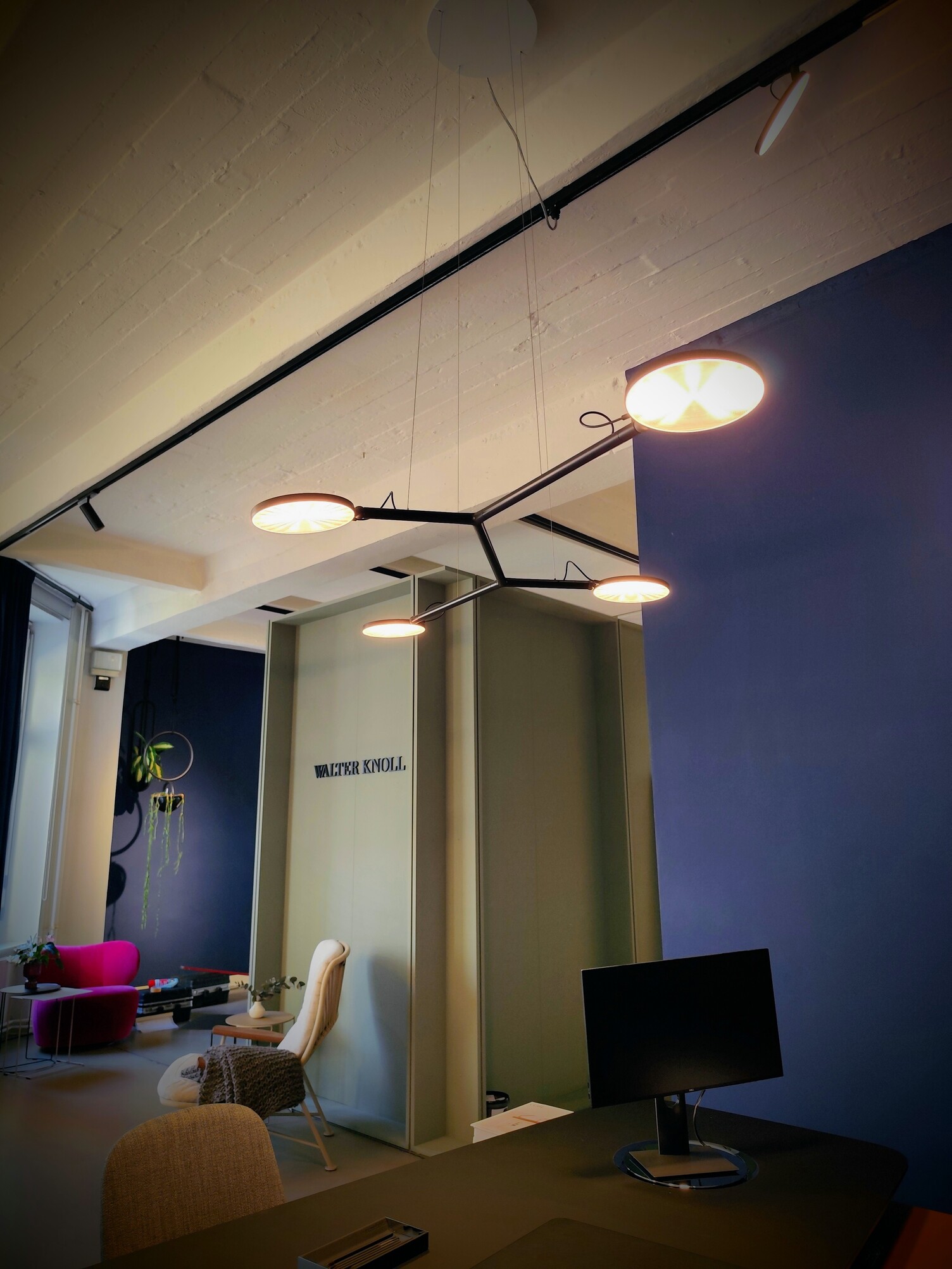 Moderne, grafische Office Hängeleuchte Blick von einem Büroeinzelarbeitsplatz in einen offenen Raum mit gemütlichen Sesseln. Über dem schwarzen Bürotisch von WALTER KNOLL sieht man an der Decke eine schwarze Hängelampe mit 4 Armen, die in 4 runde flache Lichtköpfe münden.