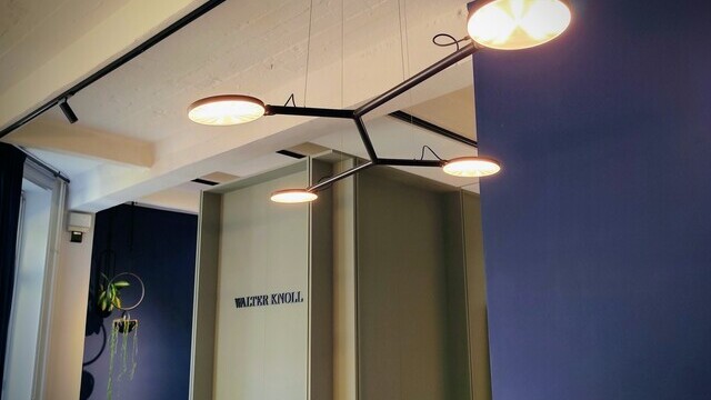 View from an individual office workstation into an open space with comfortable armchairs. Above the black office desk by WALTER KNOLL, a black pendant lamp with four arms can be seen on the ceiling, ending in four round, flat light heads.