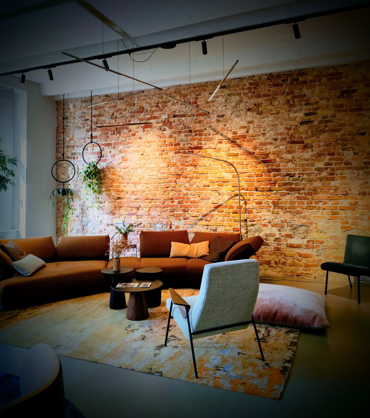 PALITO zylindrisches Stableuchtenensamble im Wohnzimmer Braune Sofalandschaft von WALTER KNOLL vor einer rötlichen Backsteinwand, direkt vor der Wand steht eine gebogene dünne Stab-Bodenlampe PALITO FLOOR. Von einer Stromschiene über den kleinen Coachtischen sind dünne waagerechte Stableuchten, PALITO, in unterschiedlichen Höhen und Winkeln abgependelt.