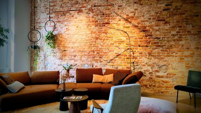 Brown sofa suite by WALTER KNOLL in front of a reddish brick wall, with a curved, slender PALITO FLOOR lamp standing directly in front of the wall. Thin horizontal PALITO lamps are suspended at different heights and angles from a power rail above the small coffee tables.