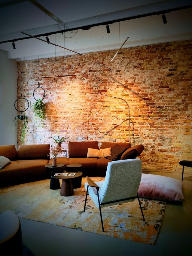 Brown sofa suite by WALTER KNOLL in front of a reddish brick wall, with a curved, slender PALITO FLOOR lamp standing directly in front of the wall. Thin horizontal PALITO lamps are suspended at different heights and angles from a power rail above the small coffee tables.