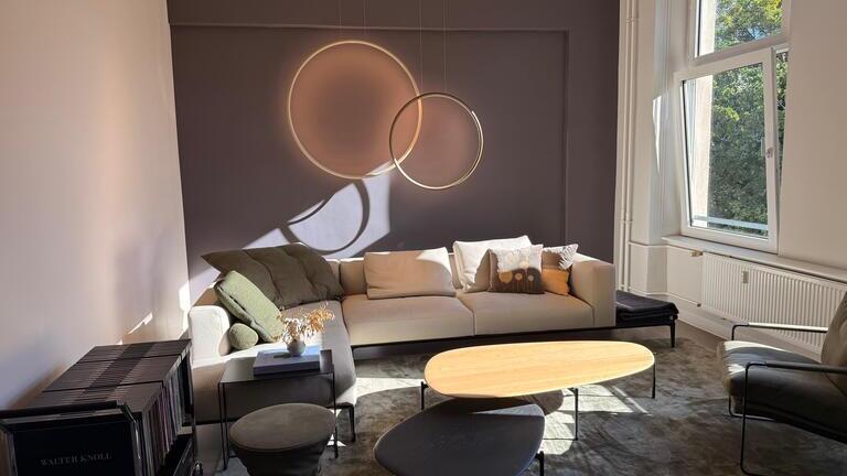 Light-coloured angular sofa suite by Walter Knoll with green and white cushions. On the grey wall behind it hang two light rings, GIRATA vertical by SATTLER, of two different diameters, which overlap.