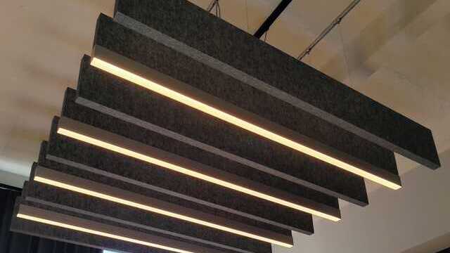 Acoustic solution. Look at the white ceiling, from which a dark grey linear acoustic baffle and an acoustic baffle with a light strip at the end are suspended in a row, alternating with each other. 