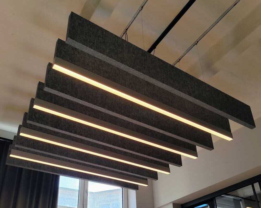 Office acoustic solution with baffles (dark grey) Acoustic solution. Look at the white ceiling, from which a dark grey linear acoustic baffle and an acoustic baffle with a light strip at the end are suspended in a row, alternating with each other.
