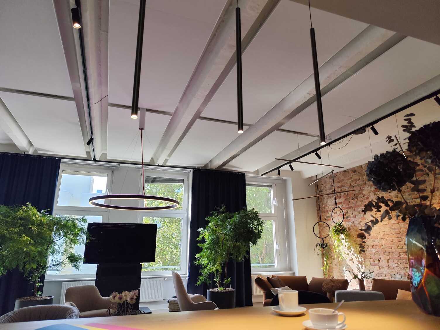 Zylindrische Pendelleuchten für die Theke Blick von der Theke aus durch einen zylindrischen Pendelleuchtenteppich aus 3 FINO PENDANT in eine Wohnlandschaft mit rundem Tisch und Stühlen über dem eine runde Ringleuchte, Doppio hängt und einer Sofalandschaft mit weiteren horizontalen Stableuchten Palito.