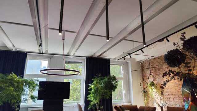 View from the bar through a cylindrical canopy of 3 FINO PENDANT lights into a living area with a round table and chairs, above which hangs a round Doppio ring light, and a sofa area with additional horizontal Palito tube lights.