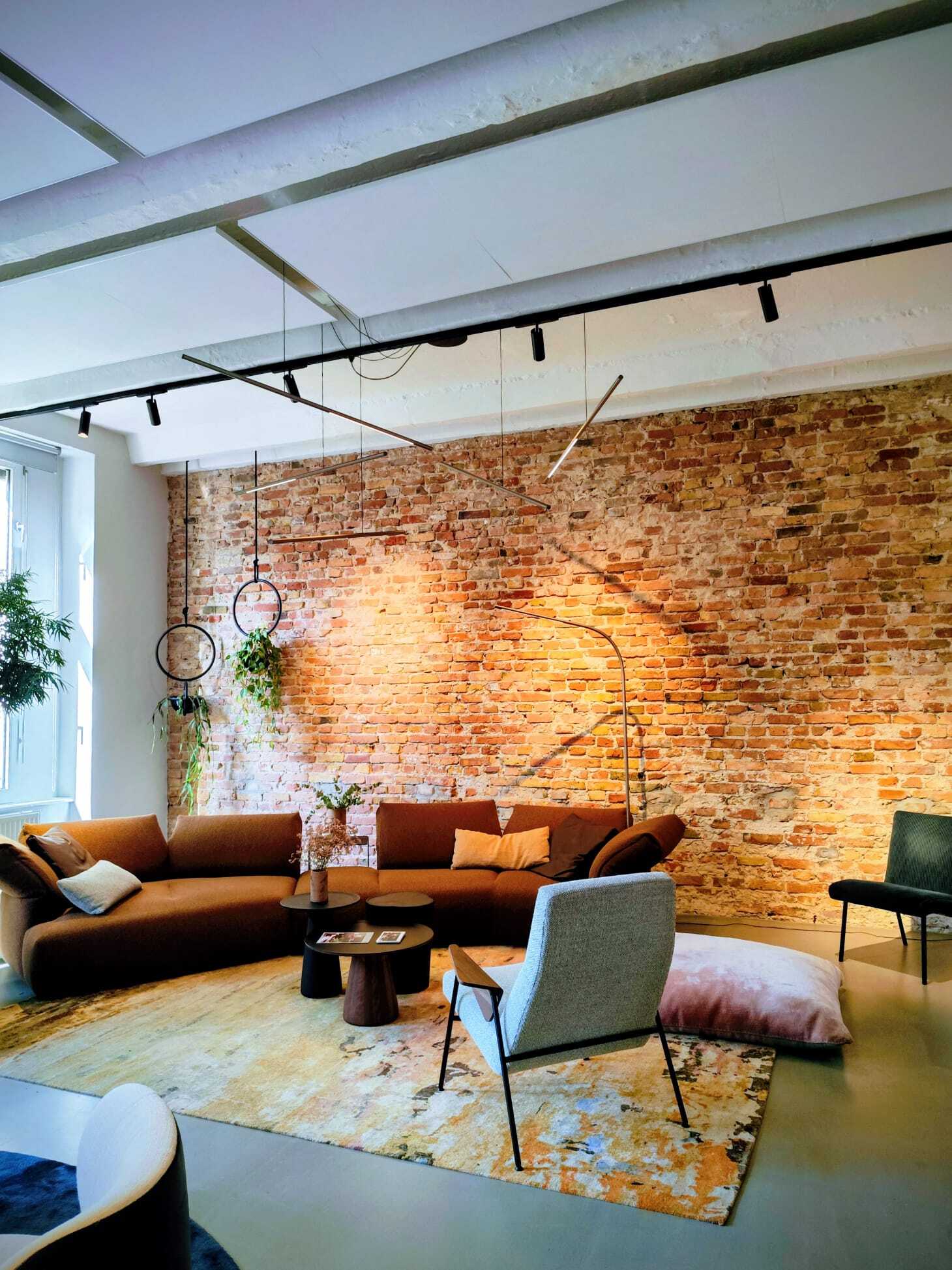 Stylische Wohnlandschaft mit Pendelleuchte PALITO Braune halbrunde Sofalandschaft und rosa Sitzkissen von WALTER KNOLL vor einer rötlichen Backsteinwand, direkt vor der Wand steht eine gebogene dünne Stab-Bodenlampe PALITO FLOOR. Von einer Stromschiene über den kleinen Coachtischen sind dünne waagerechte Stableuchten, PALITO, in unterschiedlichen Höhen und Winkeln abgependelt.