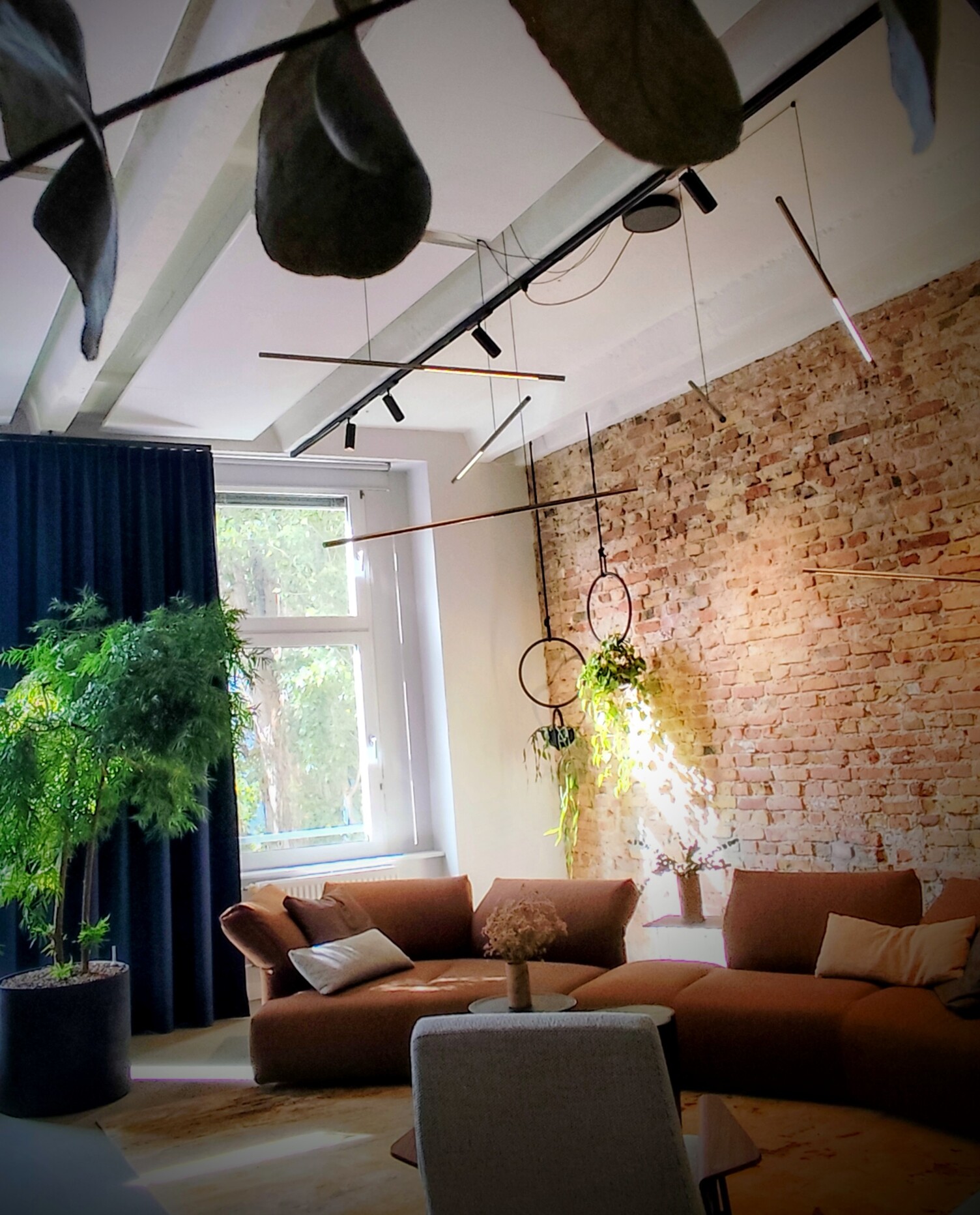 Lichtensemble für das Wohnzimmer Braune halbrunde Sofalandschaft von WALTER KNOLL vor einer rötlichen Backsteinwand. Von einer Stromschiene über den kleinen Coachtischen sind dünne waagerechte Stableuchten, PALITO, in unterschiedlichen Höhen und Winkeln abgependelt. Deckennah auch eine Stromschiene in der einzelne bewegliche zylindrische schwarze Spots EVOLUTION SPOTs positioniert sind und weitere Lichtakzente im Raum schaffen.