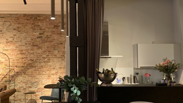 Black bar table by Walter Knoll with three narrow bronze-coloured pendant lights, Fino Pendant, along its length. In the background, a view into a small kitchen.