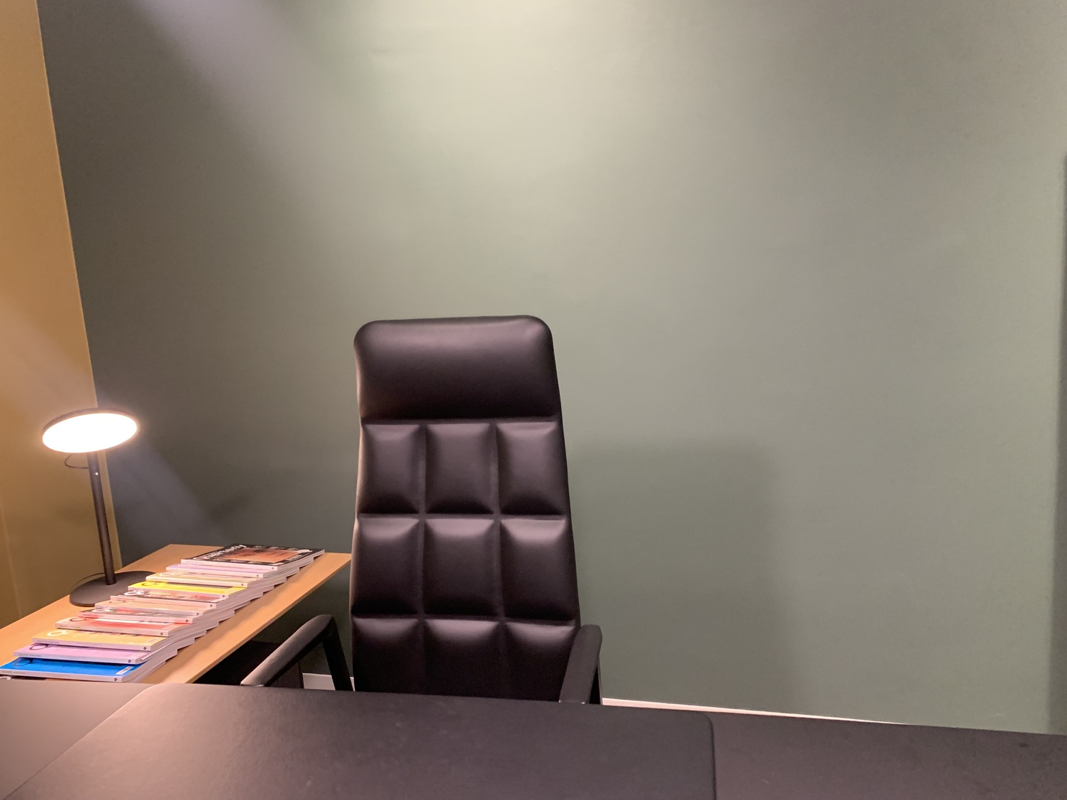Bürotischleuchte Blick auf einen Einzelarbeitsplatz mit schwarzem Tisch und schwarzem Ledersessel von Walter Knoll. Daneben eine Holz-Beistellanrichte, auf der viele Zeitschriften aufgefächert liegen, diese werden von einer schwarzen Bürotischleuchte mit großem runden flachen Lichtkopf, AVVENI TABLE, angestrahlt.