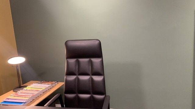 View looking at a single workstation with a black table and black leather armchair by Walter Knoll. Next to it is a wooden sideboard with lots of magazines fanned out on it, illuminated by a black desk lamp with a large round flat light head, AVVENI TABLE. 