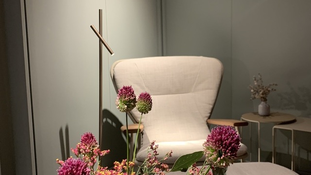 View over a purple and green bouquet of flowers onto a Shinzo lounge chair by Walter Knoll, brightly lit by a delicate cylindrical floor lamp, Fino Floor by Sattler, standing in front of a green wall with a black Walter Knoll logo.