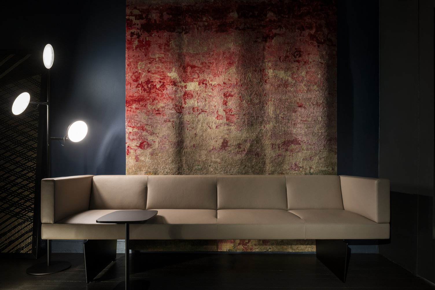 Sofa with ambient lighting from the three-headed AVVENI FLOOR 3 floor lamp Light-coloured sofa by Walter K. in front of a warm mural in shades of red hanging on a blue wall; on the left hand side stands a black floor lamp with three round luminous heads, the AVVENI FLOOR 3.