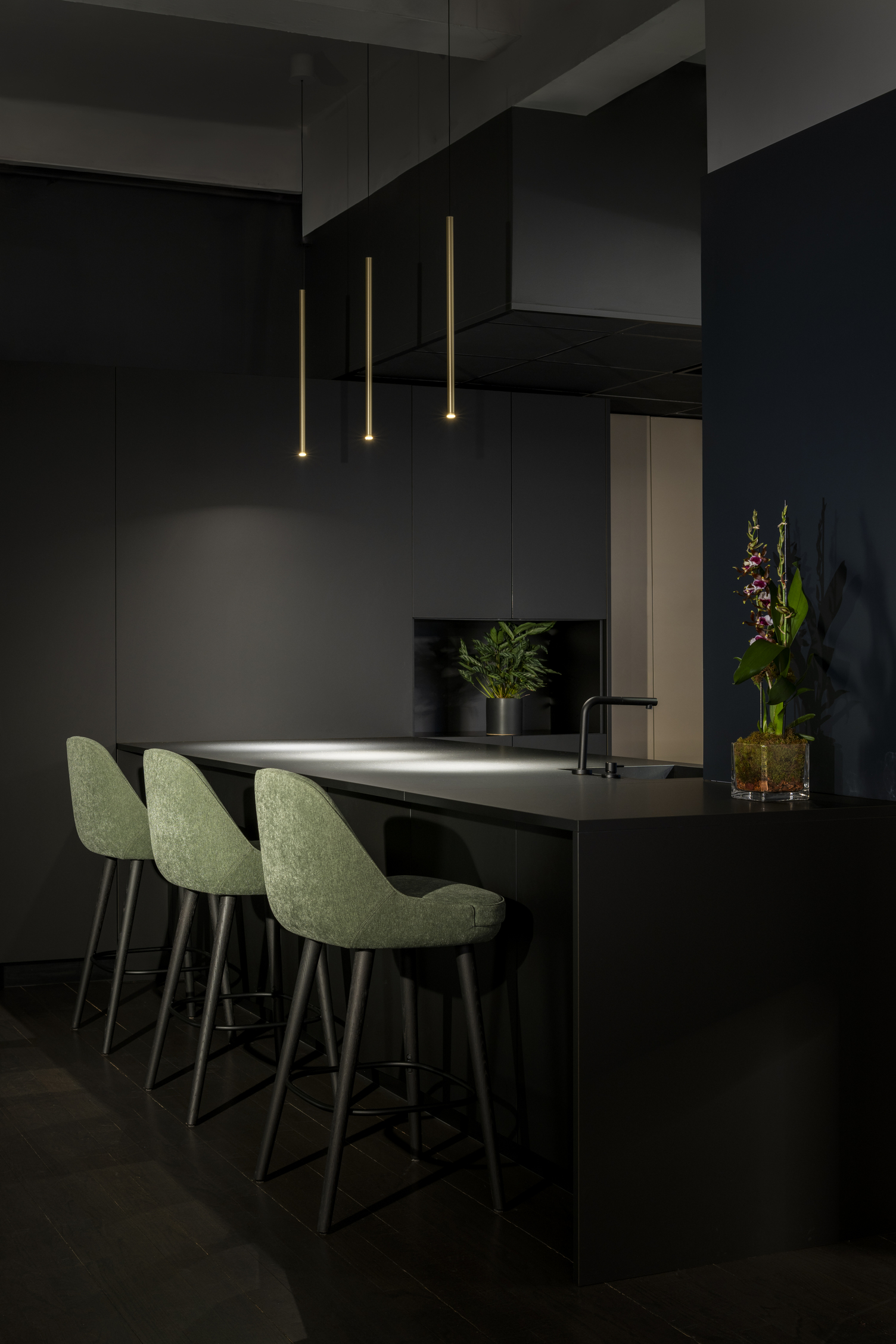 Counter with FINO PENDANT cylindrical pendant lamps. Black-grey high dining counter with green upholstered high chairs suspended above three thin, rod-shaped champagne-coloured FINO PENDANT pendant lights along the length.