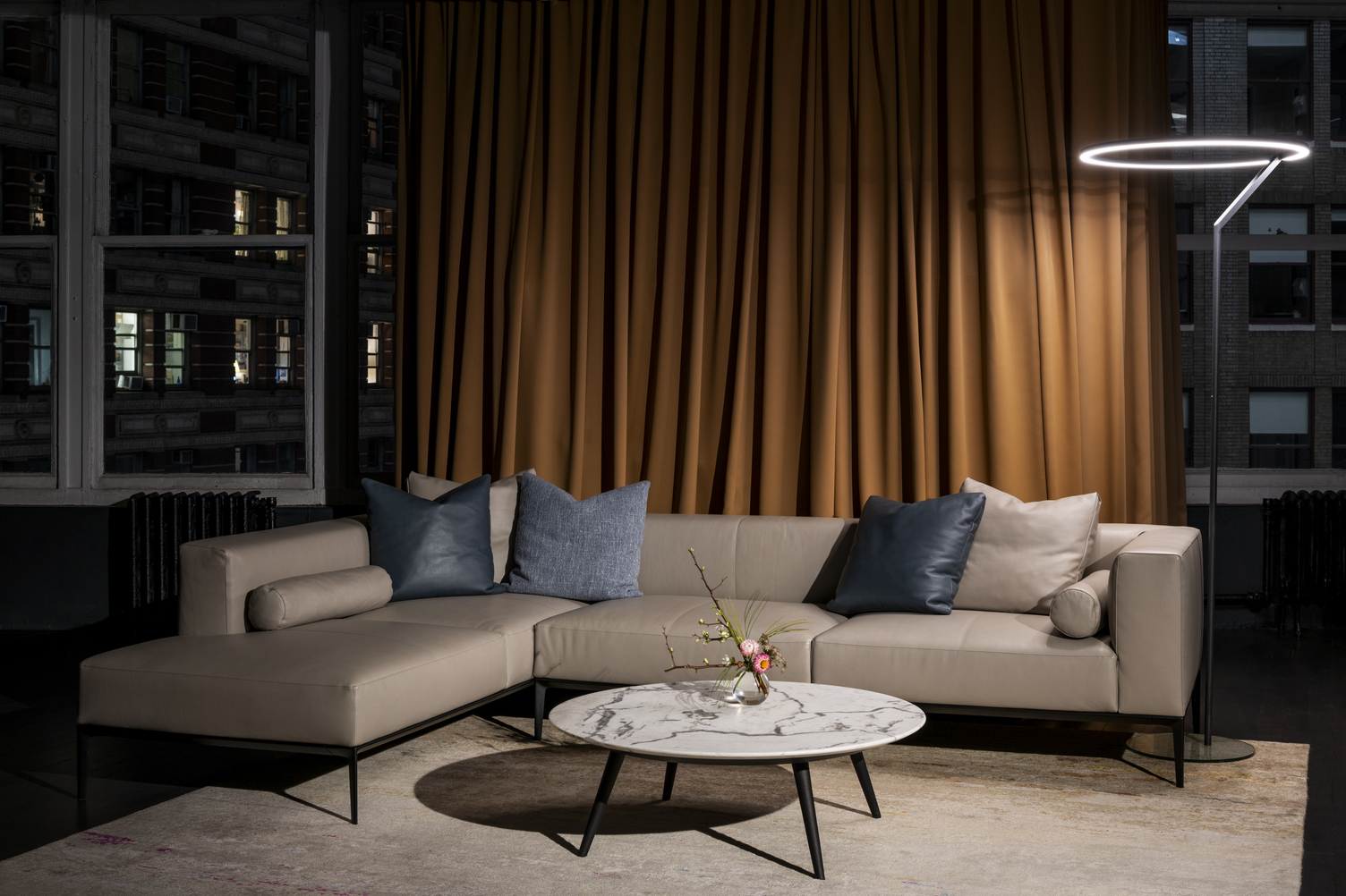 Elegant floor lamp as a ring lamp INSOSPESO FLOOR Light beige sofa by Walter K. in front of a mustard-coloured curtain. There is a blanket and two cushions in the corner of the sofa. Behind the sofa is an elegant black floor lamp, the INSOSPESO FLOOR. The cylindrical base of the floor lamp transitions into a ring light that shines directly onto the sofa.