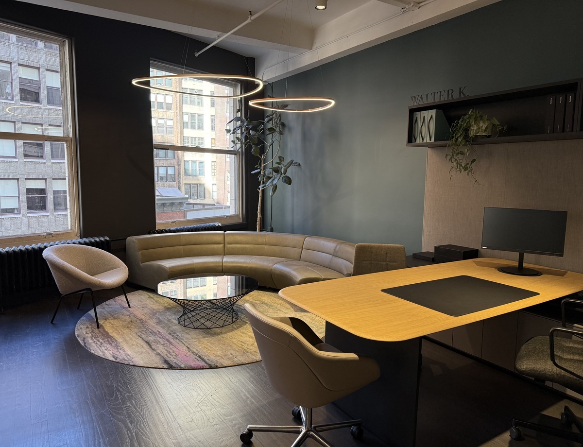 Gemütliche Besprechungsecke mit dreieckigen SLIM DELTA Pendelleuchten Blick in den Walter K. Showroom, über einen Holztisch hin zu einer Sofaecke, bestehend aus einem Sessel und viertel Kreissegment Sofa in elegantem beige, darüber sind dreickige Ringleuchten von SATTLER abgependelt.