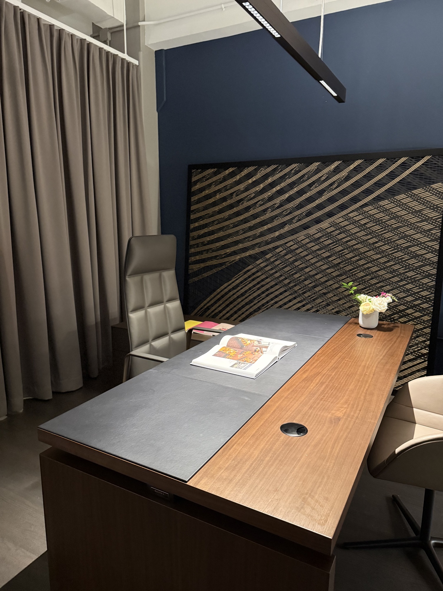 Blendfreie Linearleuchte PALADIO LENS über Fokusarbeitsplatz Holz-Schreibtisch mit Leder-Bürostuhl vor einem grauen Vorhang. Der Tisch ist hell erleuchtet durch eine lineare Arbeitsplatzleuchte PALADIO LENS, die entlang der Tischlänge abgependelt ist.