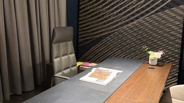 Holz-Schreibtisch mit Leder-Bürostuhl vor einem grauen Vorhang. Der Tisch ist hell erleuchtet durch eine lineare Arbeitsplatzleuchte PALADIO LENS, die entlang der Tischlänge abgependelt ist.