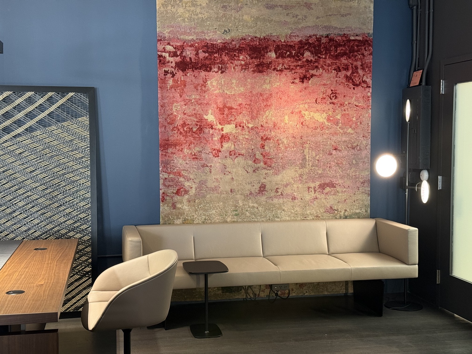 Sofa mit Ambientelicht durch 3köpfige Stehleuchte AVVENI FLOOR 3 Helles Sofa von Walter K. vor einem warmen Wandbild in Rottönen, das an einer blauen Wand hängt, in der einen Ecke steht eine schwarze Stehlampe mit 3 runden leuchtenden Köpfen, die AVVENI FLOOR 3.