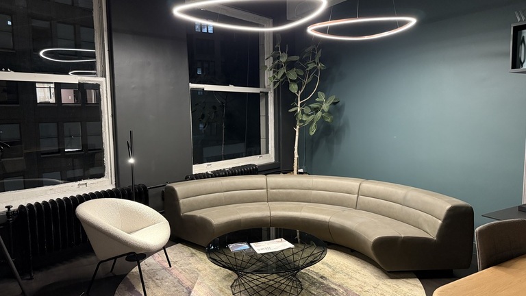 Blick nachts in den Walter K. Showroom zu einer Sofaecke von Walter Knoll, bestehend aus einem Sessel und einem Viertelkreissegment Sofa in elegantem beige, darüber sind dreickige Ringleuchten von SATTLER abgehangen.