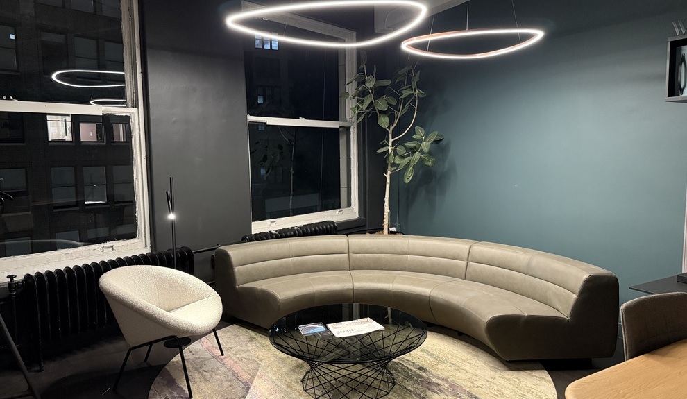 Halbrunde Sofaecke mit Pendelleuchten abgerundeter Dreiecke Blick nachts in den Walter K. Showroom zu einer Sofaecke von Walter Knoll, bestehend aus einem Sessel und einem Viertelkreissegment Sofa in elegantem beige, darüber sind dreickige Ringleuchten von SATTLER abgehangen.