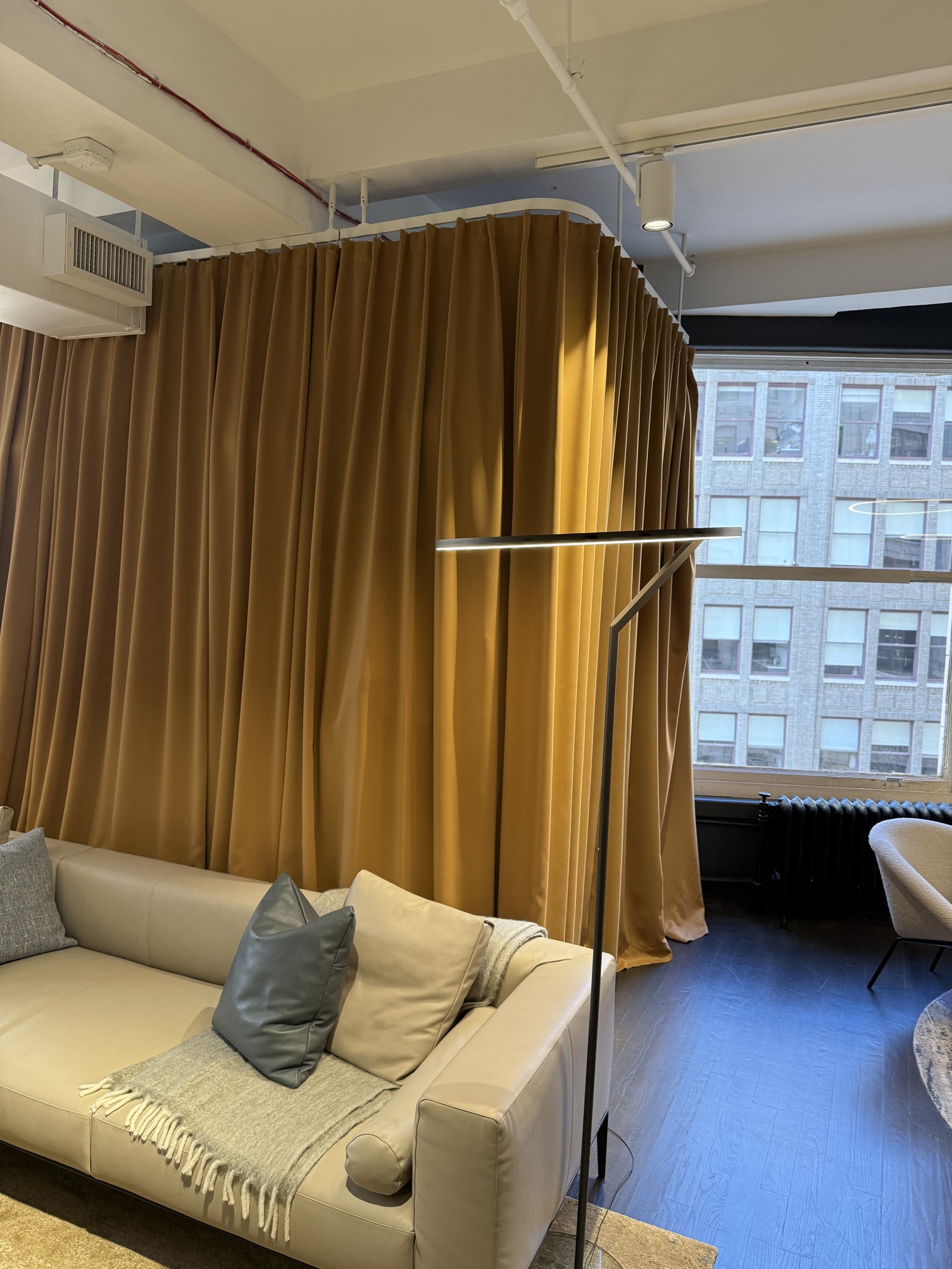 Elegante Stehleuchte als Ringleuchte INSOSPESO FLOOR Hell-Beiges Sofa von Walter K. vor einem Senffarbenen Vorhang. Im Sofaeck liegt eine Decke und zwei Kissen. Hinter dem Sofa steht eine elegante schwarze Stehleuchte, die INSOSPESO FLOOR. Der zylindrische Fuß der Stehlampe geht in ein Ringleuchte über, die direkt auf das Sofa scheint.